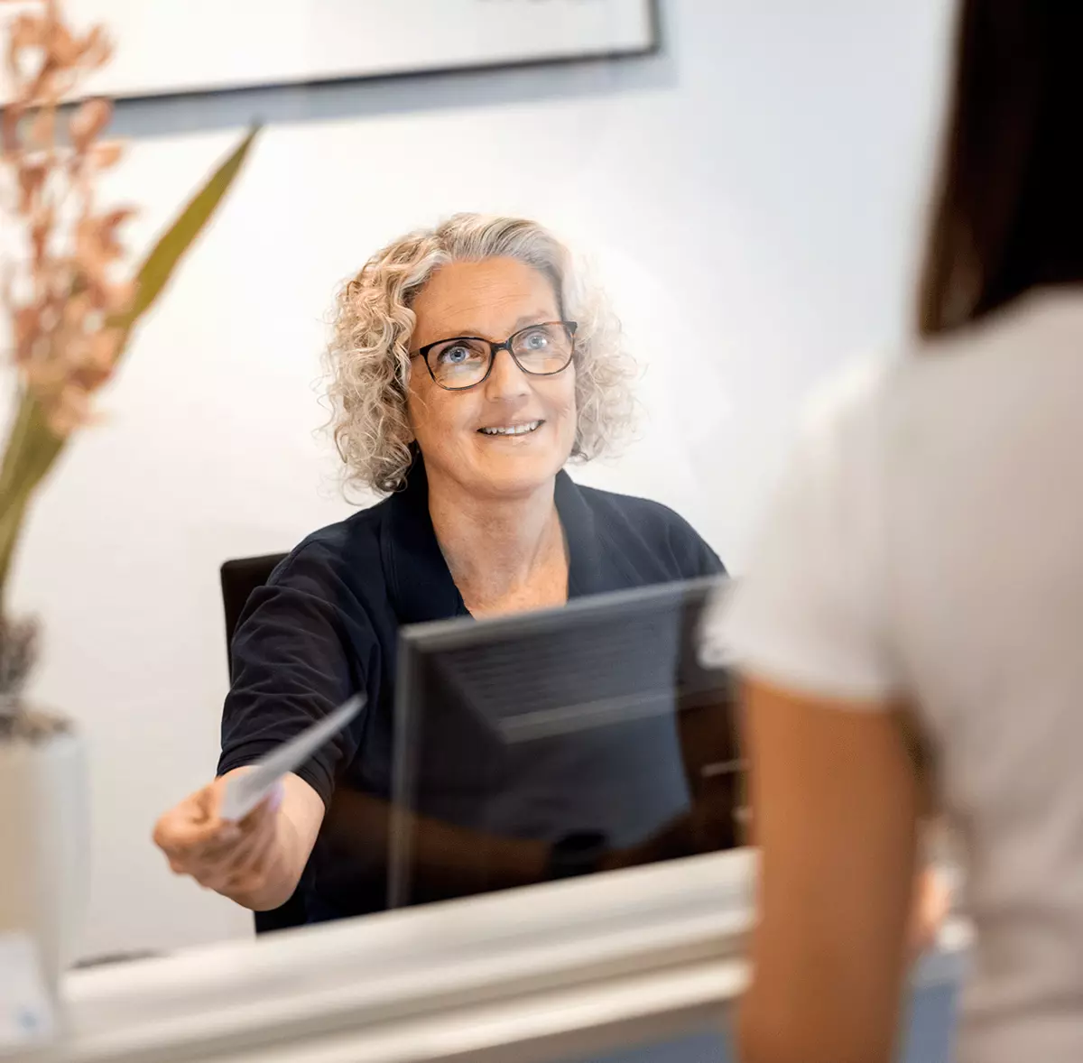 Eine freundliche Mitarbeiterin reicht einer Patientin am Empfang wichtige Informationen